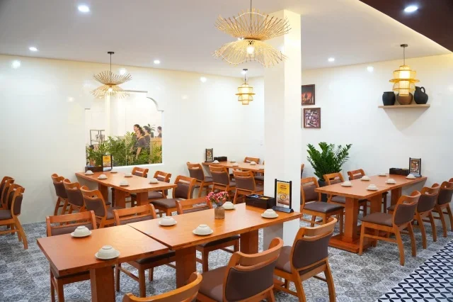 Góc bàn ăn bằng gỗ mộc mạc, ánh sáng vàng dịu, tường gạch thô và quạt trần kiểu xưa, tạo không gian ấm áp. Một gia đình đang quây quần bên mâm cơm.